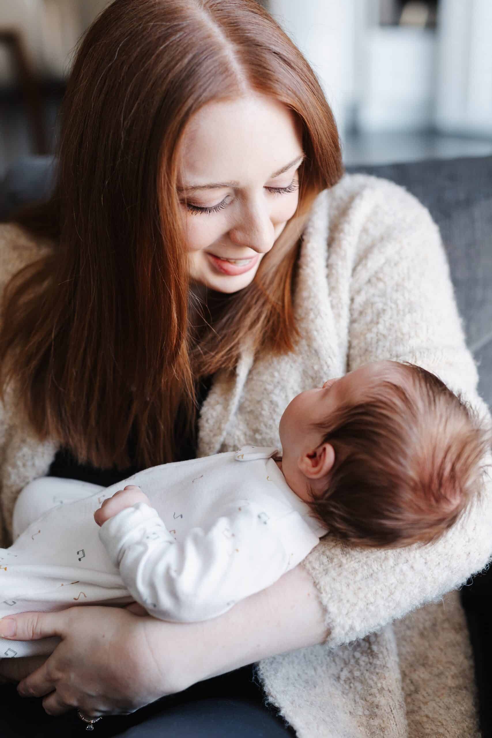 A mother in a beige sweater sits on a couch smiling down to her newborn baby in her arms