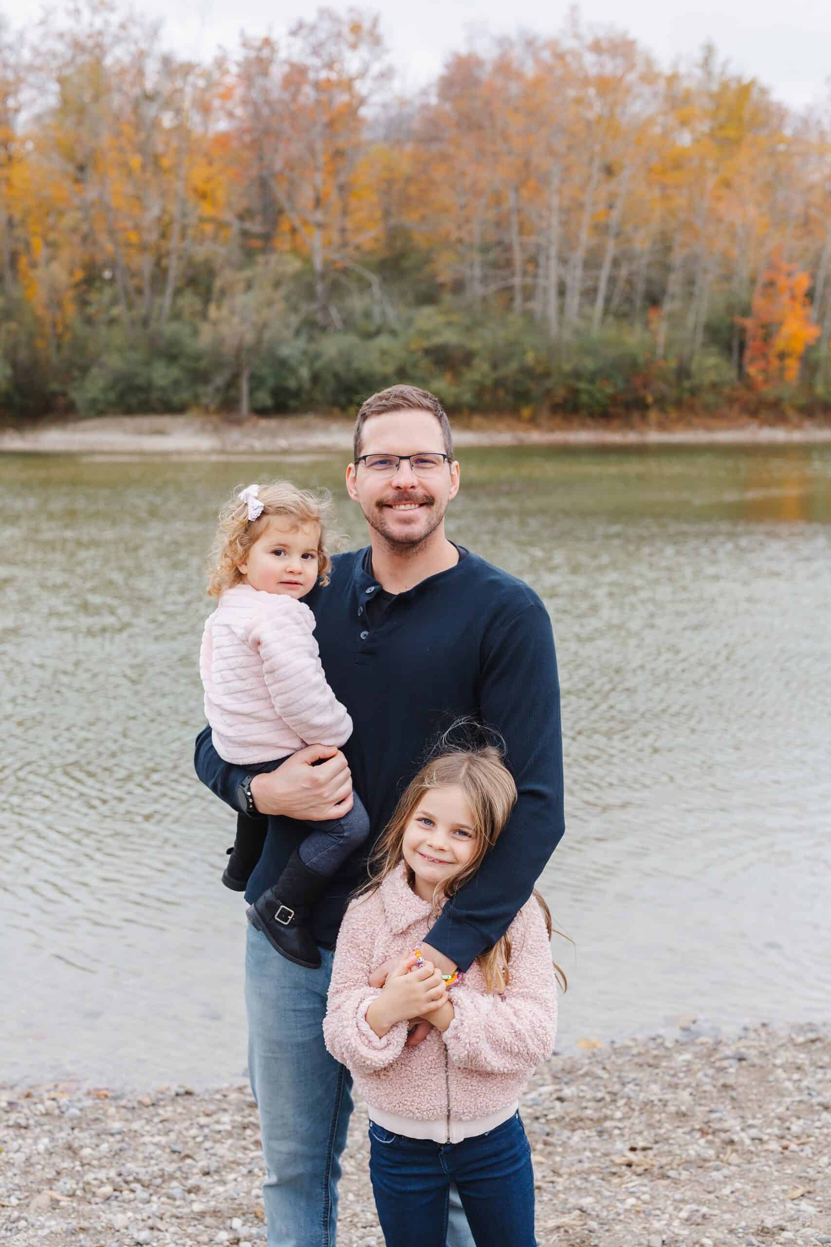 A father stands on the edge of a river in fall with his two toddler daughters in pink sweaters