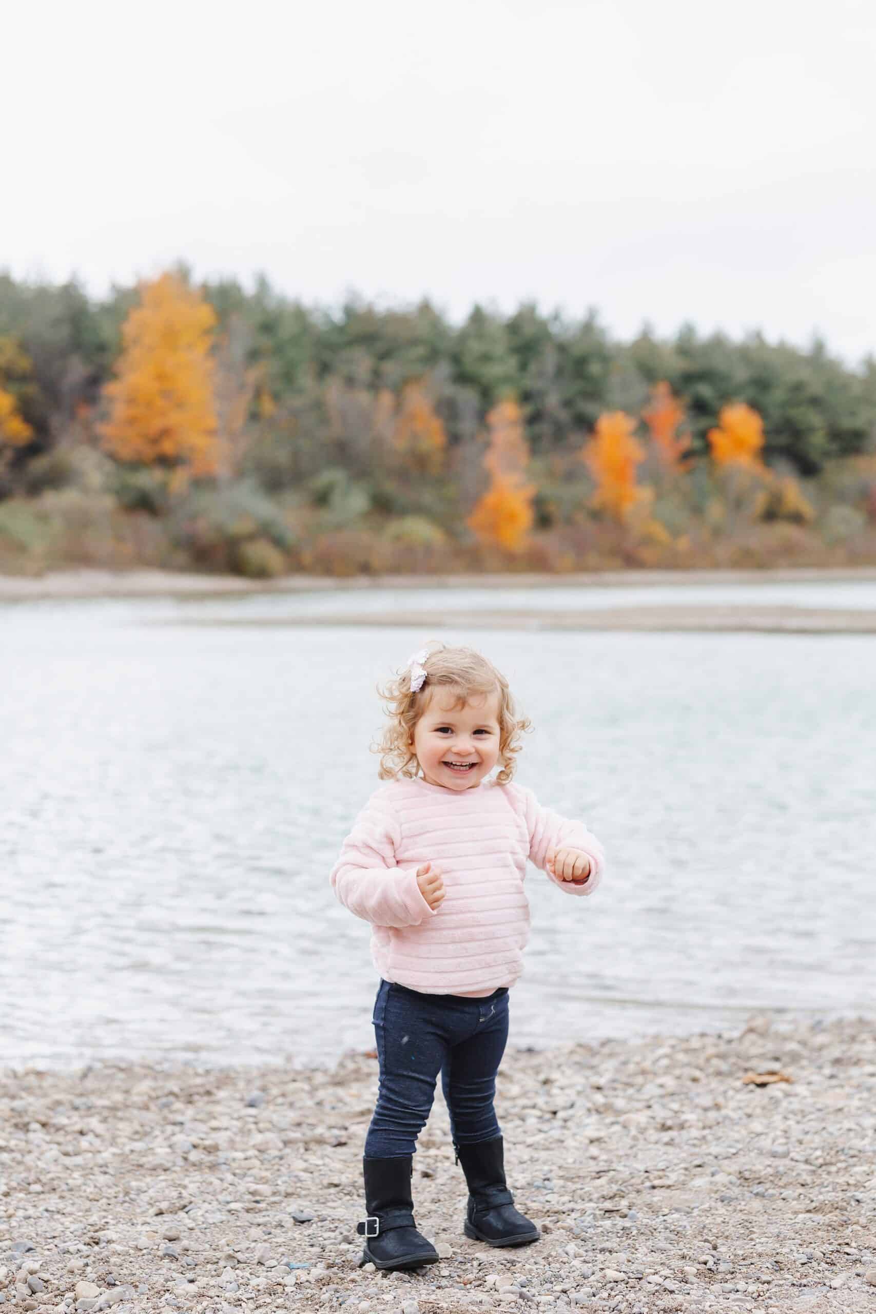 A young toddler girl in a pink sweater smiles while playing on the edge of a river before a guelph storm game