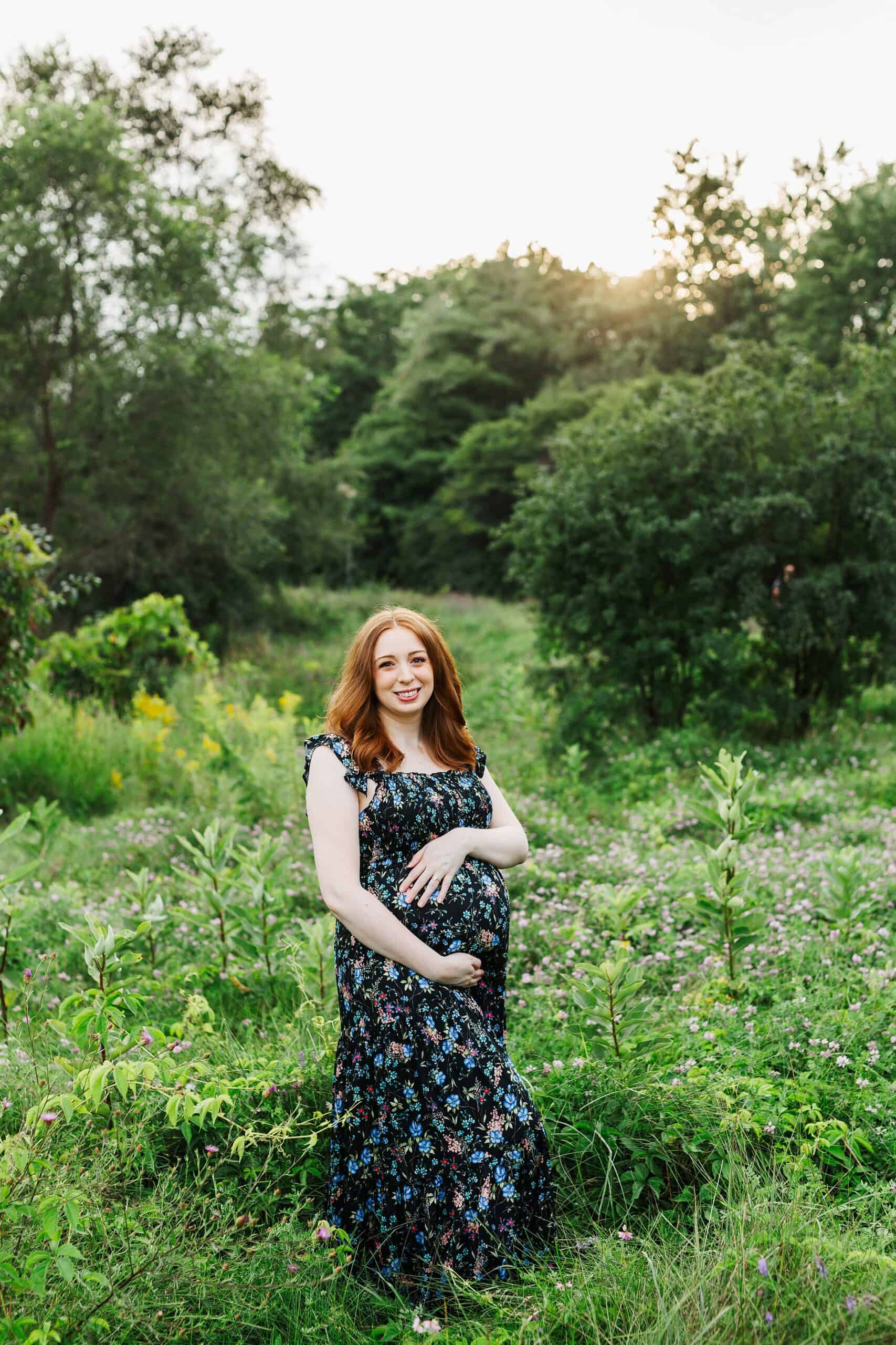 A mother to be holds her bump while standing in a forest trail at sunset in a blue maternity dress after visiting gyms in guelph