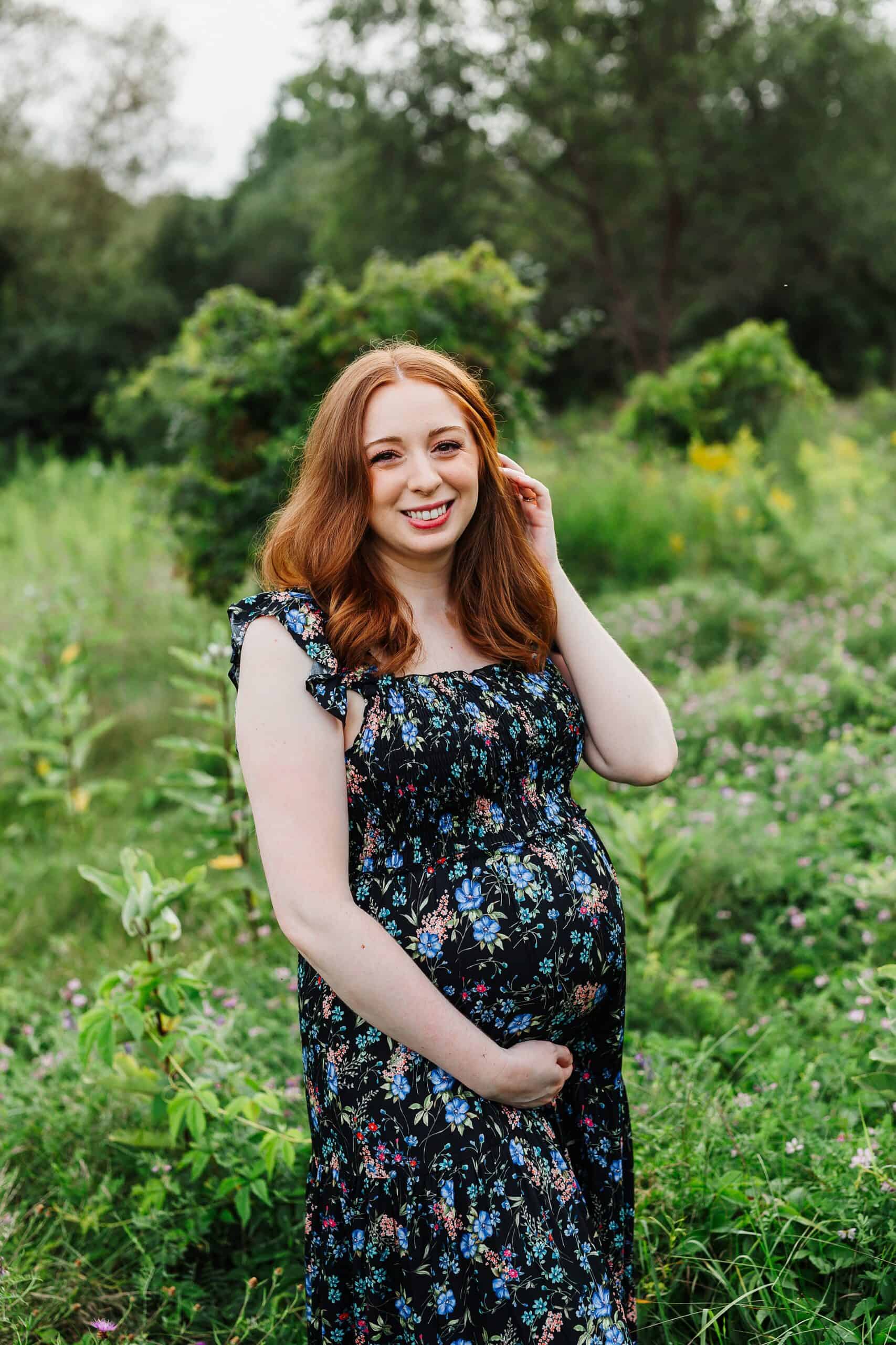 A mother to be in a blue floral print maternity dress stands in an overgrown patch of wildflowers with a hand in her hair after visiting gyms in guelph