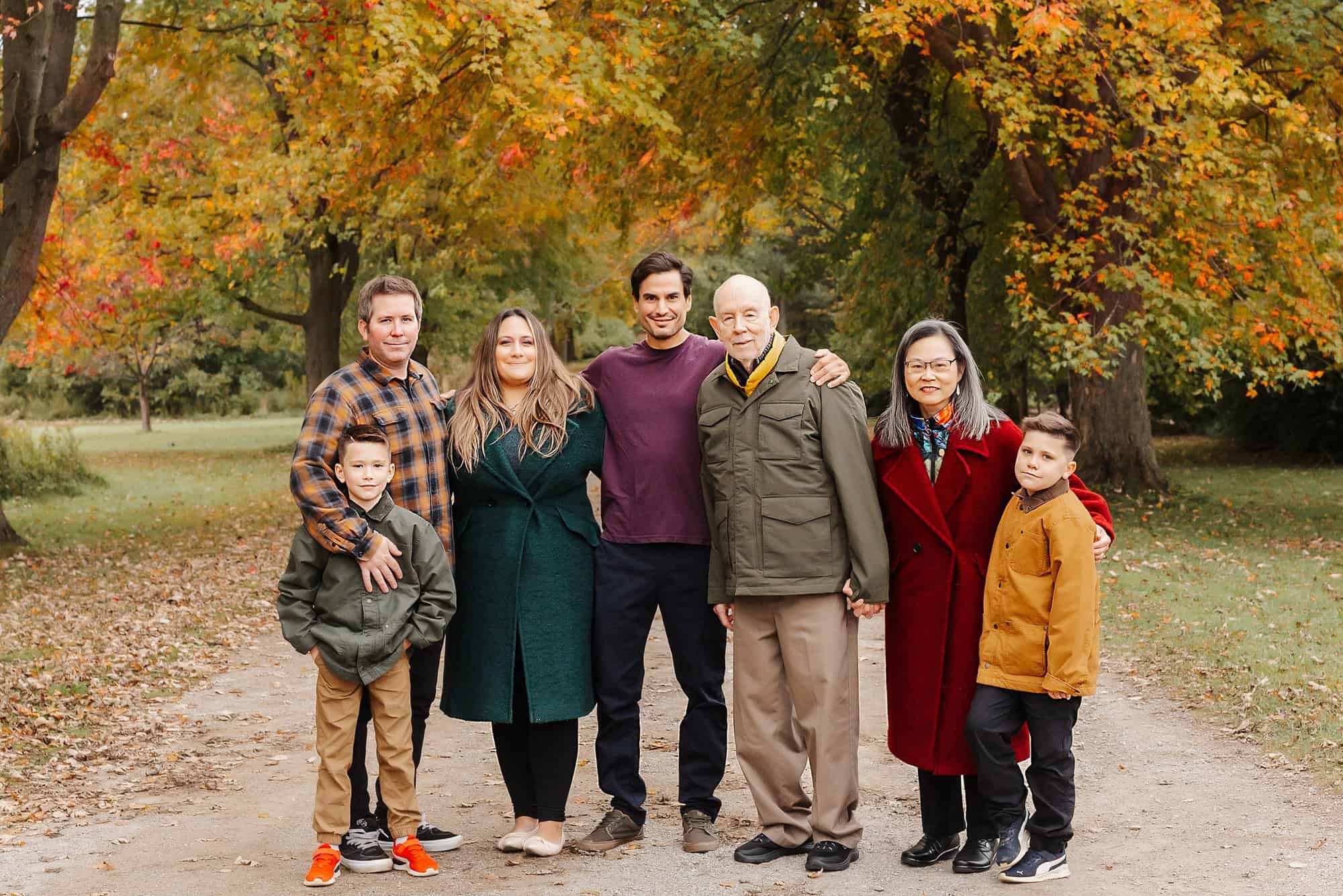 A family of 7 with three generations stands in a park path holding hands in fall before visiting dynamo gymnastics