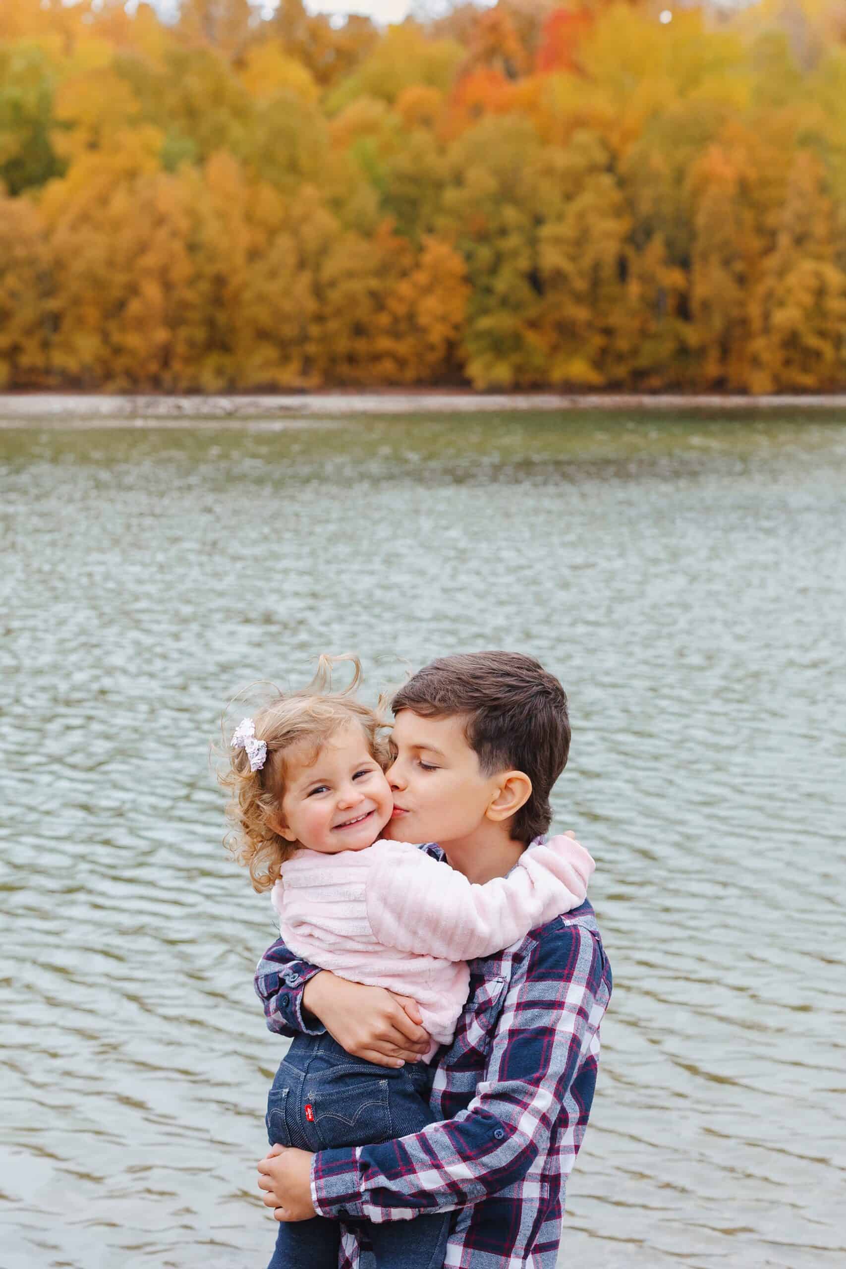 A young boy holds and kisses his toddler baby sister while standing along a lake